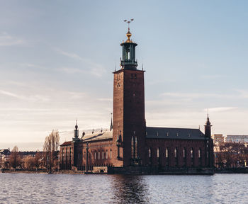 Lighthouse by river amidst buildings against sky