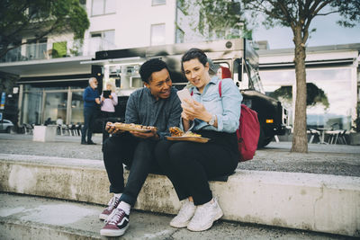 Smiling friends sharing smart phone while sitting with meal against food truck in city