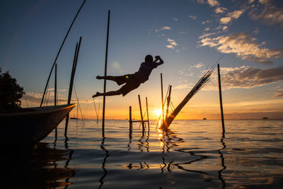 Silhouette boy jumping from boat in sea against sky during sunset