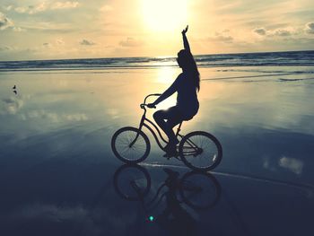 Woman riding bicycle at beach against sky during sunset