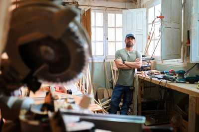 Self assured male woodworker with folded arms and beard looking at camera against instruments in workroom