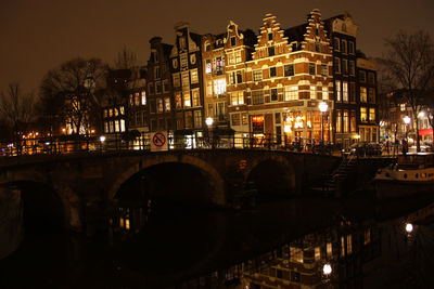 Illuminated bridge over river by buildings against sky at night