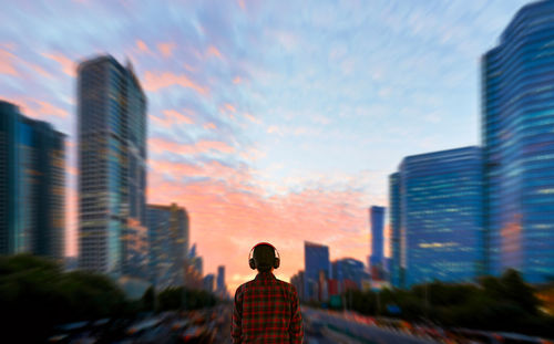 Rear view of man standing by buildings against sky during sunset