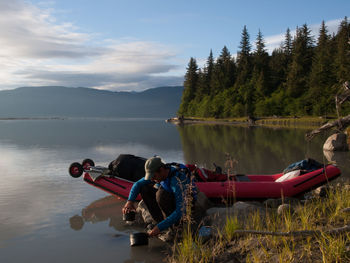 Man filling water in container while crouching by boat at lakeshore against trees