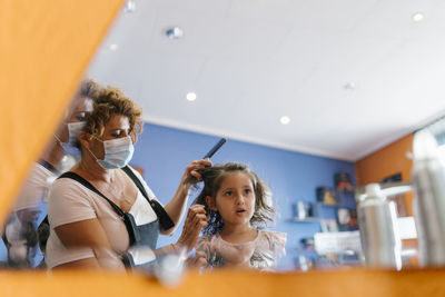 Mature female hairdresser cutting girl's hair seen from mirror reflection at barber shop