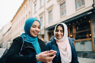 Smiling young woman sharing mobile phone with female friend walking in city