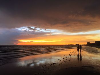 Silhouette friends walking on beach against cloudy sky during sunset