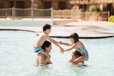 Side view of cheerful little boy and girl in swimwear holding hands and laughing while sitting on shoulders of mother and older sister standing in outdoor swimming pool in sunlight