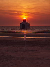 Silhouette of a wooden house on the beach with the sun sitting on the chimney during sunset