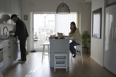 Businesswoman talking on smart phone while boyfriend doing chores in kitchen