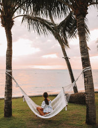 Woman sitting on hammock at beach against cloudy sky during sunset