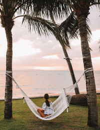 Man sitting on hammock by tree against sky