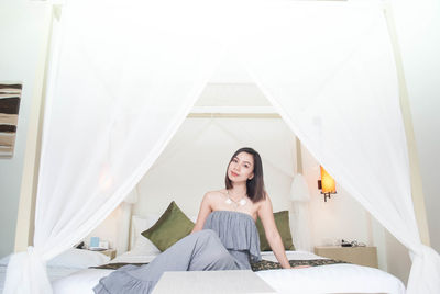 Portrait of beautiful young woman in off shoulder dress sitting on bed at home