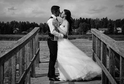 Side view of newlywed couple romancing while standing on pier against sky