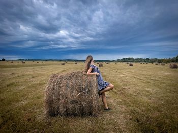 Woman in dress standing on a field near a bale of hay and looking at the dark clouds in the sky