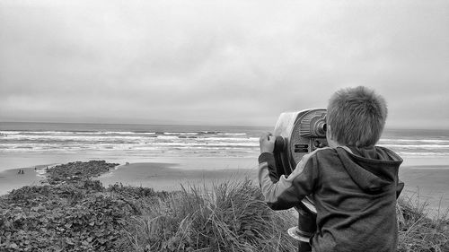 Rear view of boy looking through coin-operated binoculars at beach against sky