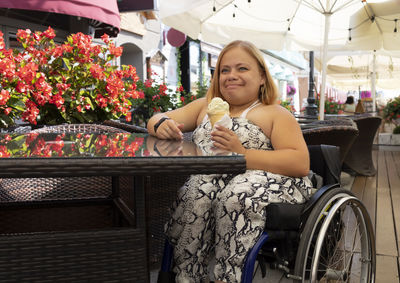 Young woman with short stature on wheelchair eats ice-cream in cafe. female adult with physical