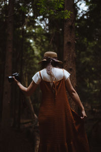 Rear view of woman holding camera while standing in woodland