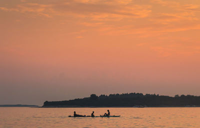 Silhouette people on paddleboard in sea against orange sky