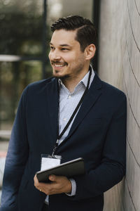 Smiling male entrepreneur standing with digital tablet leaning on wall looking away