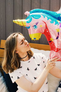 Smiling woman with closed eyes holding unicorn balloon against wall