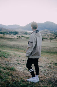 Back view of young man in casual outfit standing on rocky cliff and admiring incredible view against cloudless sky during sunset