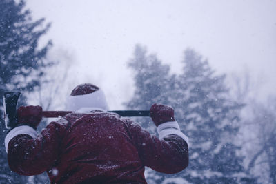 Rear view of mid adult man in santa costume holding axe while standing in forest during snowfall