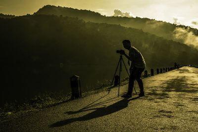 Full length of man standing by tripod on road by lake during sunset