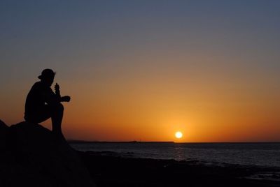 Silhouette man sitting at beach against sky during sunset