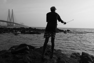 Rear view of man fishing while standing on rock at beach against sky