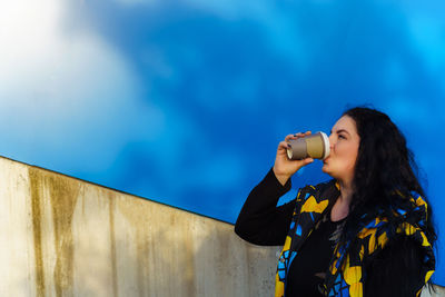 Brunette woman  drinks coffee from a paper cup on a background of blue wall