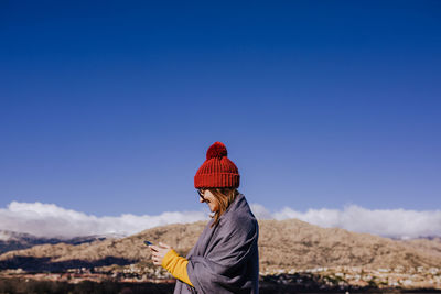 Side view of woman using phone while standing against mountain