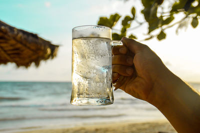 Close-up of hand holding beer glass against sea and sky