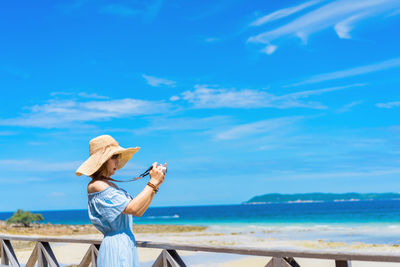 Side view of woman photographing at beach against blue sky