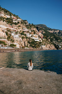 Rear view of woman sitting on rock by sea against sky