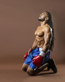 Full length of shirtless boxer kneeling on brown background