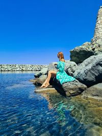 Woman sitting  on rock by sea against clear blue sky