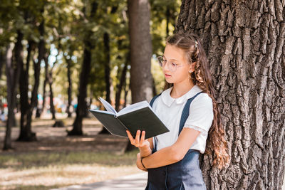 Schoolgirl in glasses reads a book near a tree in the park.