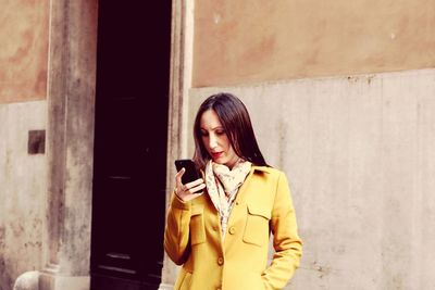 Young woman using phone while standing against wall