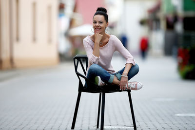 Full length portrait of smiling woman sitting on chair in city