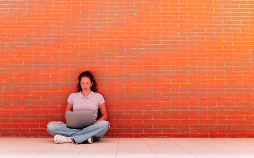 Full body of focused female student in casual wear surfing internet on netbook while sitting on floor near brick wall