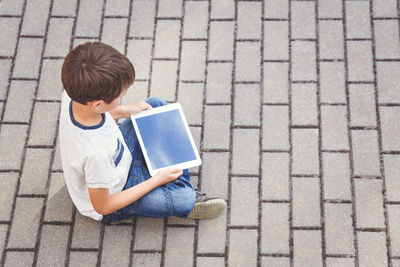 High angle view of boy holding digital tablet while sitting on footpath