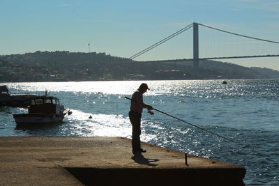 Side view of man fishing at harbor with bosphorus bridge in background
