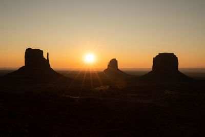 The sun rises over the horizon as a car passes in front of the mittens in monument valley.