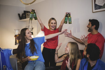 Cheerful woman holding beer bottles standing with friends in living room