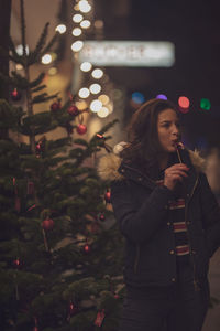 Woman eating food while standing against christmas tree at night