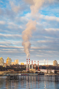 Smoke emitting from factory by river against sky