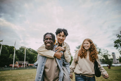 Teenage girl looking at braided hair boy piggybacking male friend at park