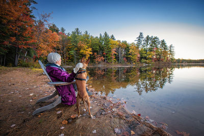 Woman with dog sitting by lake on chair during autumn