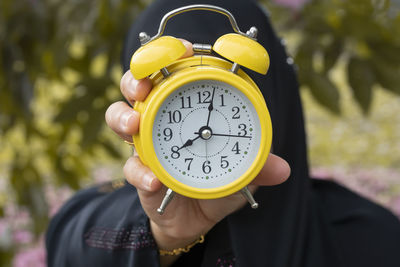 Close-up of person holding yellow alarm clock against trees